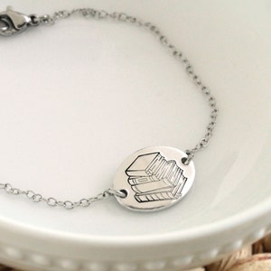 May include: A silver chain bracelet with a round charm featuring a stack of books. The charm is engraved with a detailed outline of the books.