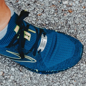 Puede incluir: Primer plano de una zapatilla de running azul con dise&ntilde;o negro y amarillo. La zapatilla tiene una etiqueta met&aacute;lica unida a los cordones que dice "You've got this".