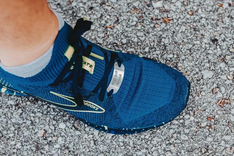 Puede incluir: Primer plano de una zapatilla de running azul con dise&ntilde;o negro y amarillo. La zapatilla tiene una etiqueta met&aacute;lica unida a los cordones que dice "You've got this".