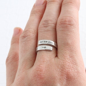 May include: A silver ring with the words "Let Him Lead" engraved on it. The ring is wrapped around a finger and features a small cross on the band.