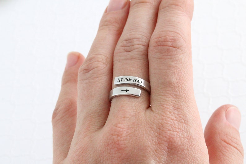 May include: A silver ring with the words "Let Him Lead" engraved on it. The ring is wrapped around a finger and features a small cross on the band.