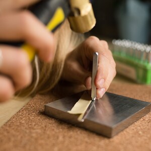 May include: A person using a metal stamp to create a design on a piece of leather. The leather is placed on a metal surface and the person is holding the stamp with their right hand. A hammer is visible in the background.