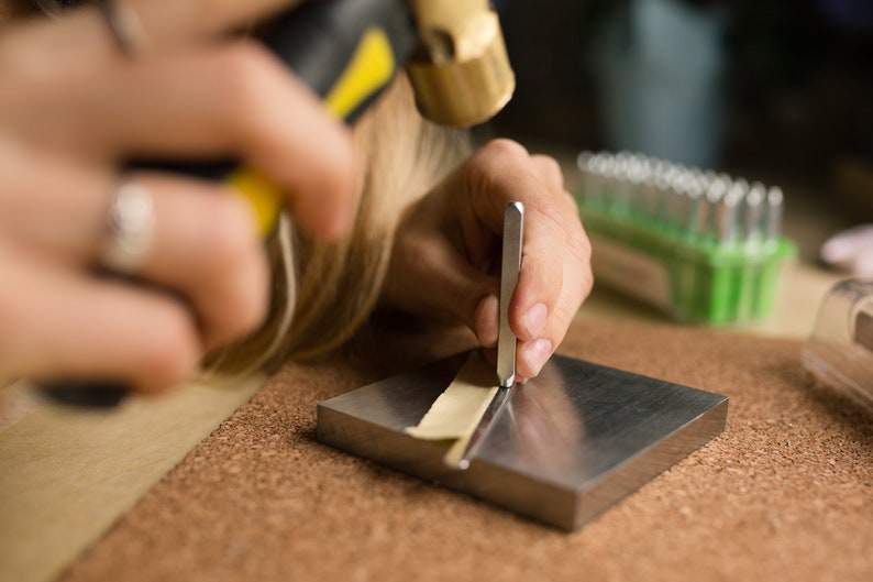 May include: A person using a metal stamp to create a design on a piece of leather. The leather is placed on a metal surface and the person is holding the stamp with their right hand. A hammer is visible in the background.