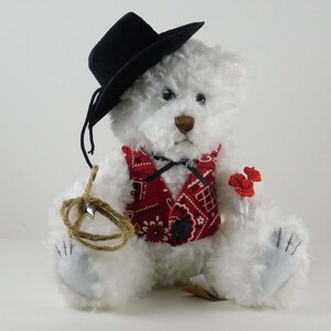 May include: White teddy bear wearing a black cowboy hat and a red bandana vest. The bear is holding a bouquet of red roses and a rope.