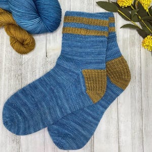 Può includere: Un paio di calzini blu lavorati a maglia con dettagli color oro sul polsino e sul tallone. I calzini sono realizzati con un filato morbido e strutturato e sono esposti su una superficie di legno chiaro. Due matasse di filato blu e oro sono sullo sfondo.