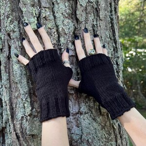 May include: A pair of black fingerless gloves made of a knitted material. The gloves are shown against a textured tree trunk background. The gloves have a ribbed cuff and cover the hand up to the knuckles.