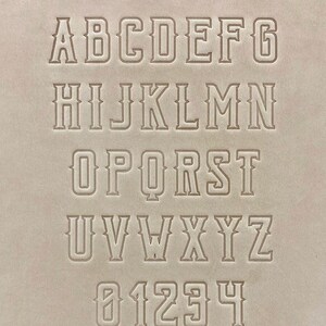 May include: A set of uppercase letters and numbers in a bold, blocky font style. The letters and numbers are outlined in a thin line and are set against a light brown background.