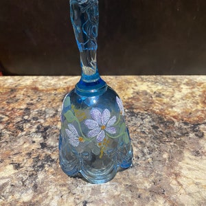 May include: A translucent light blue glass bell featuring a hand-painted floral design with glittery purple petals, green leaves, and small yellow accents. The bell has a scalloped bottom edge and a textured, sculpted handle with a clear glass finish.