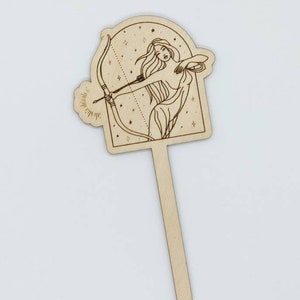May include: Wooden cake topper featuring a line drawing of a woman with long hair holding a bow and arrow, set against a starry background. The topper is shaped like an arch and has a wooden stick attached to the bottom.