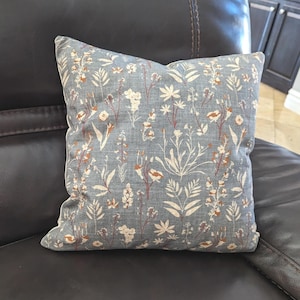 May include: Square throw pillow with a floral pattern. The pillow features a light blue background with a repeating design of white, brown, and rust-colored flowers and foliage. The pillow is placed on a dark brown leather couch.