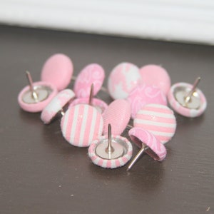 May include: A collection of pink and white patterned push pins with silver metal heads. The push pins are arranged in a loose pile on a brown surface.
