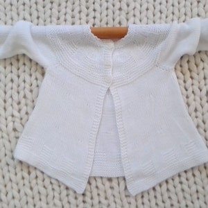 May include: White knitted baby cardigan with a button closure in the back.