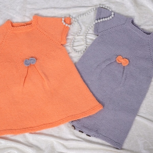 May include: Two knitted baby dresses, one in peach and one in lavender, with a bow detail on each.