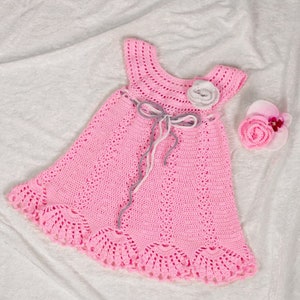 May include: A handmade pink crochet dress with a scalloped hem and a matching flower hair accessory. The dress features a decorative bow at the waist and a flower accent on the shoulder. The dress is made of soft yarn.