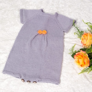 May include: A knitted baby onesie in a light purple color with orange buttons. The onesie has short sleeves and a round neckline.