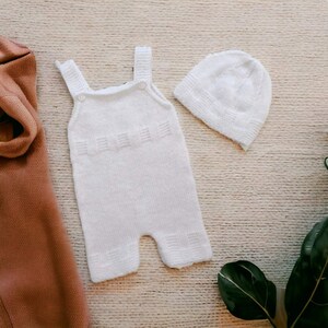 Könnte beinhalten: Weißes gestricktes Baby-Outfit mit passender Mütze. Das Outfit ist ein ärmelloser Strampler mit Knopfverschluss an der Schulter. Die Mütze ist ein einfacher Beanie-Stil.