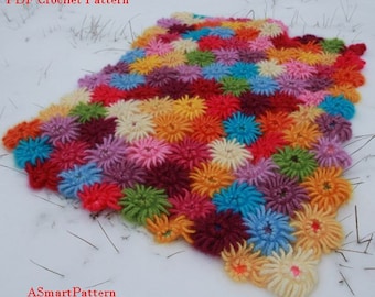 May include: A colorful crocheted blanket with a variety of flower shapes in shades of pink, red, orange, yellow, green, blue, and purple. The blanket is laid out on a white surface with a few pine needles scattered around it. The text "PDF Crochet Pattern" and "ASmartPattern" are visible in the image.