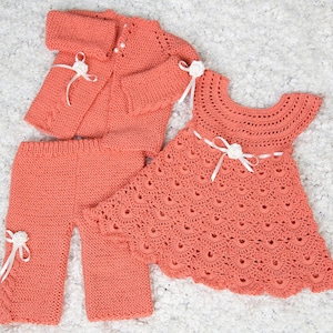 May include: A set of two crocheted baby outfits in a peach color. One outfit is a dress with a scalloped hem and the other is a cardigan and pants.