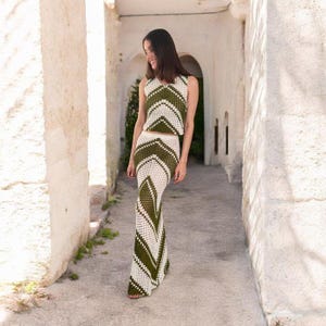May include: A two-piece crochet outfit in white and olive green. The sleeveless top and floor-length skirt feature a chevron pattern. The outfit is made of a breathable, open-weave material, perfect for warm weather. The outfit is a stylish choice for a summer event.