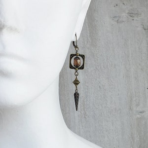 May include: Bronze earrings with a geometric square design featuring a brown faceted gemstone. The earrings dangle with a small round bead and a pointed teardrop shape.