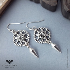 May include: A pair of silver earrings. Each earring has a decorative square design with an ornate pattern and a dangling, pointed, geometric charm. The earrings are displayed on a light blue surface.