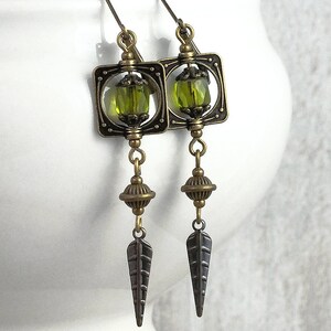 May include: A pair of bronze earrings with green glass beads and a leaf-shaped design. The earrings are hanging from a white surface.