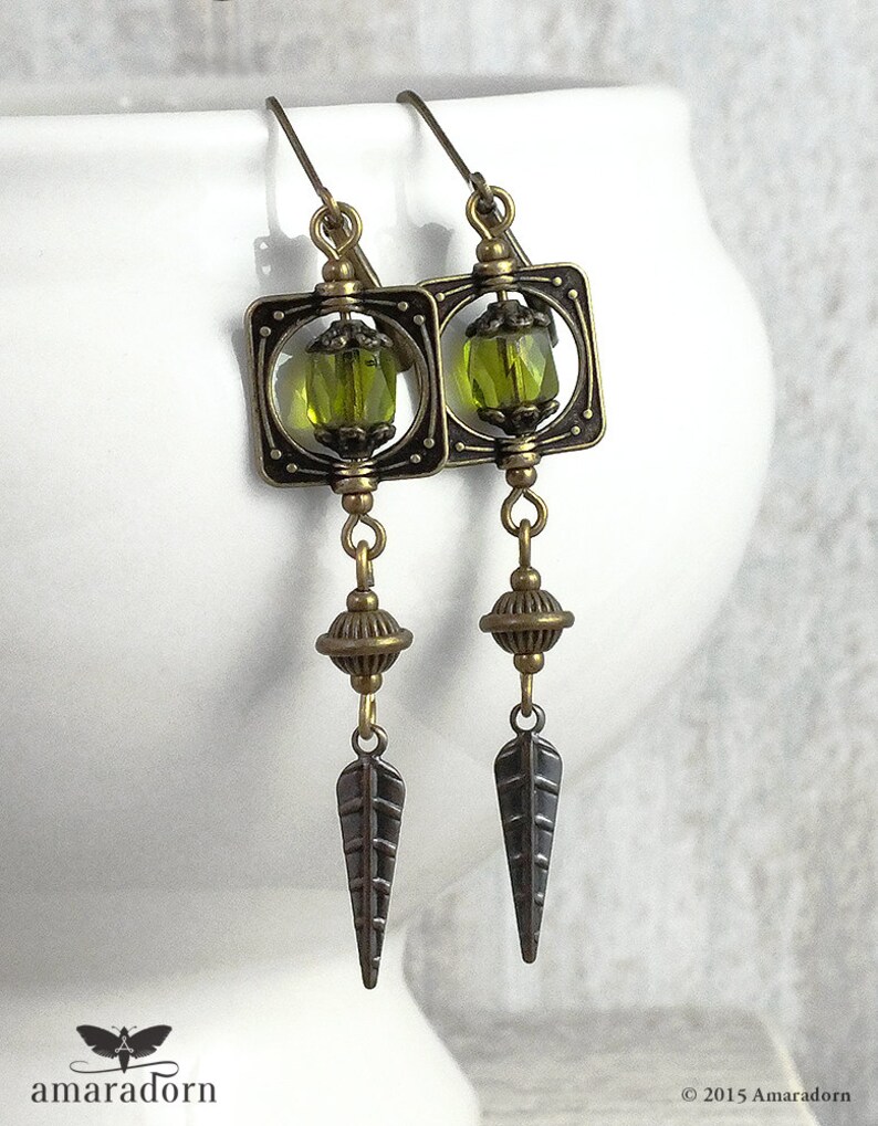 May include: A pair of bronze earrings with green glass beads and a leaf-shaped design. The earrings are hanging from a white surface.