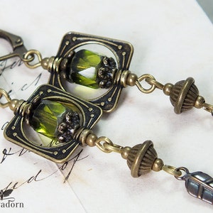 May include: A pair of green and bronze earrings with a square design and a green glass bead. The earrings have a feather charm and a small round bead.