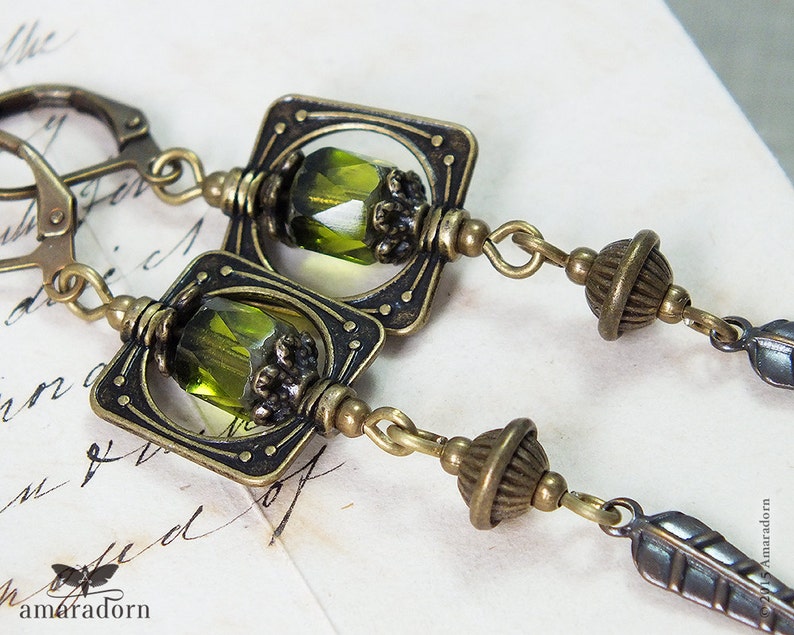 May include: A pair of green and bronze earrings with a square design and a green glass bead. The earrings have a feather charm and a small round bead.