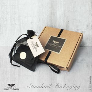 May include: A brown cardboard box with a black ribbon and a black and white logo that says "amaradorn handmade jewellery". The box is sitting on a white surface. The box is surrounded by a black and white fabric bag with a white tag that says "amaradorn".