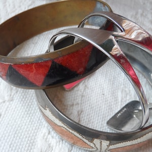 Puede incluir: Tres brazaletes de metal, uno con un patrón geométrico rojo y negro, otro con un acabado plateado y otro con un patrón de cobre y blanco.