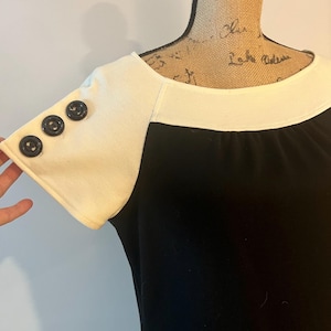 Puede incluir: Un vestido blanco y negro con cuello blanco y mangas cortas. Las mangas tienen tres botones negros a cada lado.