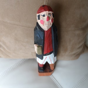 May include: Hand-carved wooden figurine of a person wearing a red hat, black jacket, red shirt, and white pants. The figurine is holding a small object in its right hand.