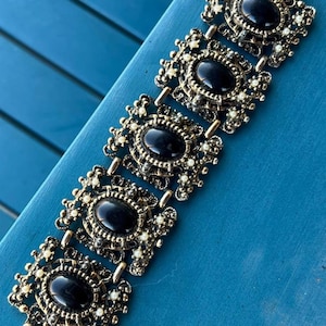 Könnte beinhalten: Ein Vintage-Armband in Goldoptik mit fünf ovalen, schwarzen Steinen. Jeder Stein ist von einem dekorativen Filigranmuster und kleinen, weißen Kunstperlen umgeben. Das Armband liegt auf einer blauen Oberfläche.