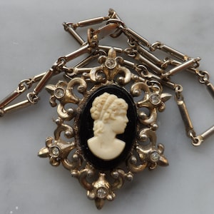 Puede incluir: Un collar plateado con un colgante de camafeo. El camafeo presenta un perfil blanco de una mujer sobre un fondo negro. El colgante está rodeado por un diseño decorativo de filigrana con diamantes de imitación.