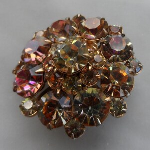 Puede incluir: Un grupo de piedras de imitación iridiscentes en tonos rosa, naranja y dorado, engastadas en una base de metal dorado. Las piedras de imitación están dispuestas en un patrón circular, creando un diseño brillante y llamativo.