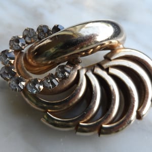Puede incluir: Un broche de tono dorado con un diseño circular que presenta un grupo de diamantes de imitación transparentes. El broche tiene una forma única y abstracta con múltiples elementos curvos.