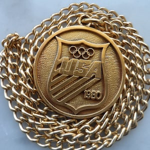 May include: Gold-colored pendant necklace with a round medallion featuring a shield with the Olympic rings and the text "USA 1980".