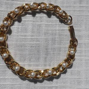 Puede incluir: Un brazalete de cadena dorada con perlas blancas falsas. El brazalete tiene un cierre de broche.