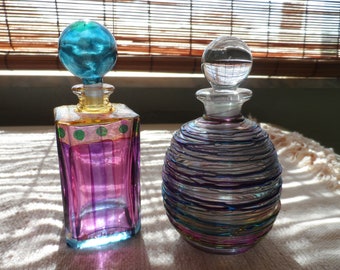May include: Two colorful glass bottles with decorative stoppers. The bottle on the left is square and has a purple, pink, and blue design. The bottle on the right is round and has a swirling design with blue, purple, and green accents.