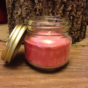 May include: A red candle in a clear glass jar with a gold lid. The candle is sitting on a wooden surface.