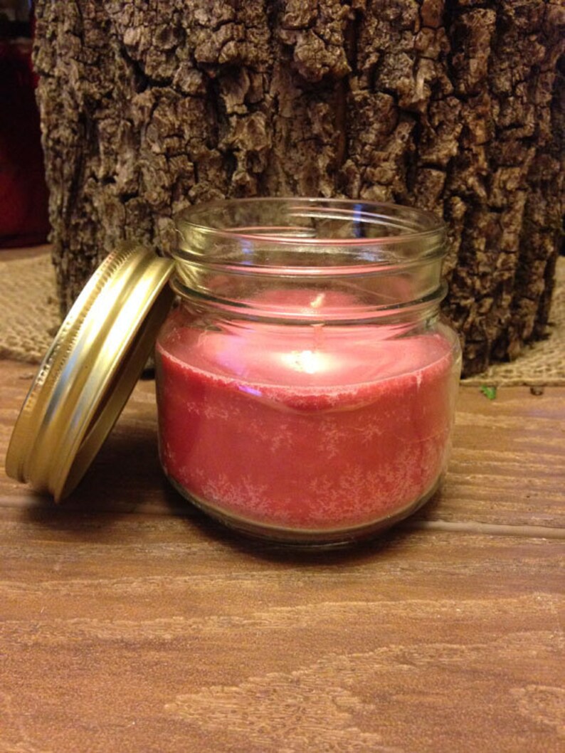 May include: A red candle in a clear glass jar with a gold lid. The candle is sitting on a wooden surface.