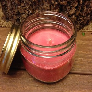 May include: A pink candle in a clear glass jar with a gold lid. The candle is sitting on a wooden surface.