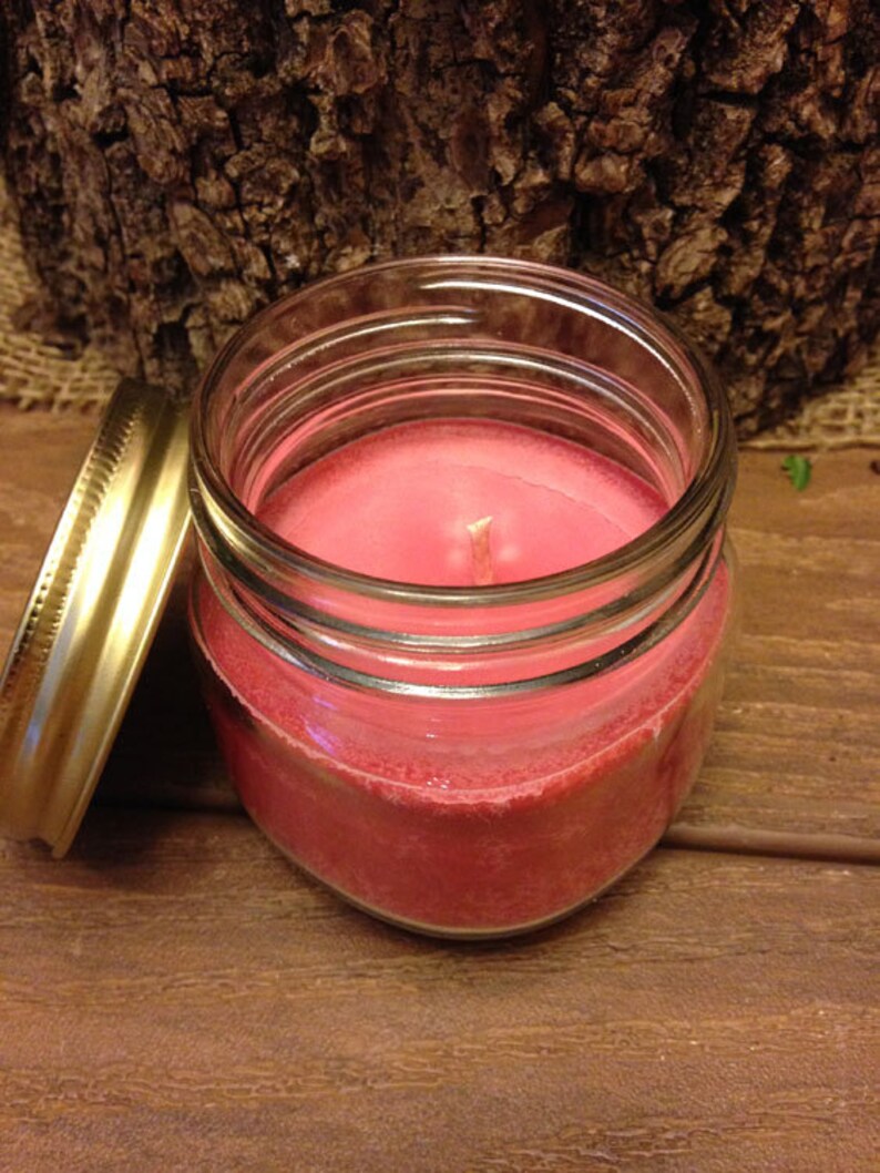 May include: A pink candle in a clear glass jar with a gold lid. The candle is sitting on a wooden surface.