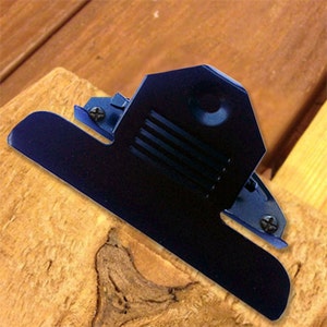 May include: A black metal clipboard with a spring-loaded clip. The clip is open and the clipboard is resting on a piece of wood.