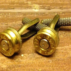May include: A pair of gold cufflinks made from repurposed bullet casings. The cufflinks have a textured design and are connected by a metal bar.