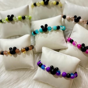 May include: A collection of colorful beaded bracelets with black Mickey Mouse charms. The bracelets are displayed on white cushions.