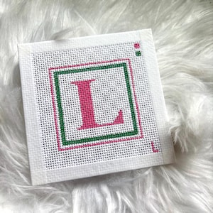 May include: A square white canvas featuring a cross-stitch design of a large pink letter L centered within a green and pink square border. Two small colored squares appear in the top right corner, and a small pink letter L is in the bottom right.
