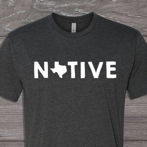 May include: A dark heather grey t-shirt with a white outline of the state of Texas and the word "NATIVE" printed on the front.