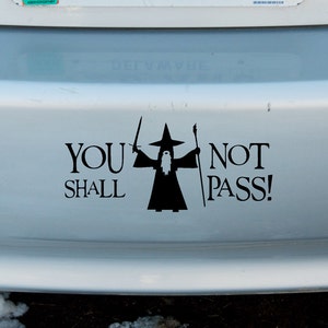 May include: A white car bumper sticker with a black silhouette of a wizard holding a sword and staff. The text reads "You Shall Not Pass!"